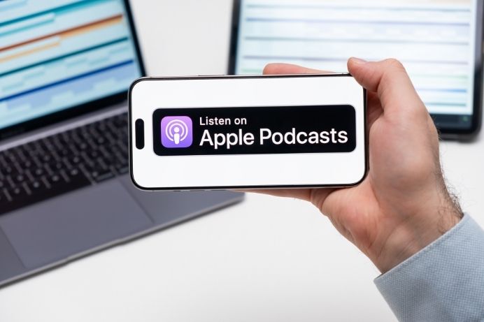 Persona sosteniendo un iPhone con la insignia "Listen on Apple Podcasts" frente a una laptop
