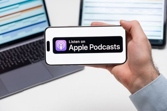 Persona sosteniendo un iPhone con la insignia "Listen on Apple Podcasts" frente a una laptop
